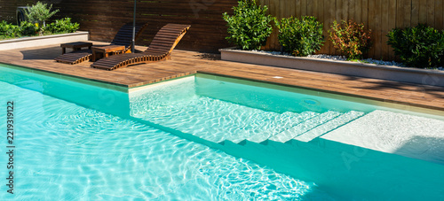 Modern swimming pool with turquoise water, wooden deck, and relaxing lounge chairs under sunny sky