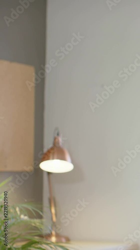 Vertical video: Sliding camera panning left to center lit copper lamp at home desk, plant entering