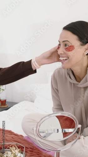 Vertical video: Smiling woman in tan hoodie having friend tuck hair on bed, with patches