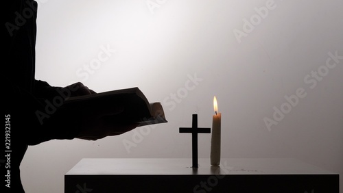 Silhouetted figure reading a sacred book beside a glowing candle and wooden cross, evoking quiet faith, prayerful devotion, spiritual reflection and serene worship
