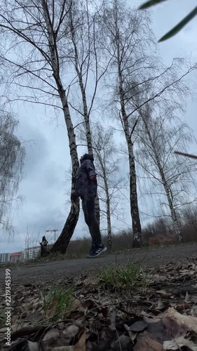 Man bending to pick object from ground under birch, hoodie and careful motion, leaf litter foreground, cloudy horizon, vertical stock video suited for action cutaway and tutorial