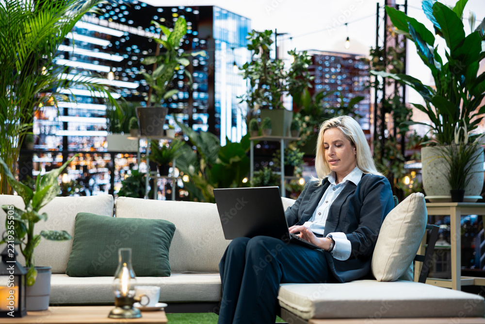 Fototapeta premium Professional woman in business attire sits with an open laptop on a rooftop lounge sofa, typing on the device for remote work from a business terrace with an urban skyline setting above the city.