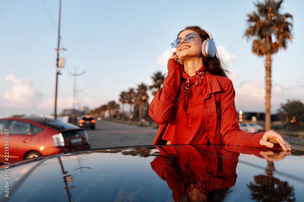 Naklejka premium Smiling woman wearing a red coat and headphones leaning on a glossy car hood outdoors with palm trees in the background. Fashion street style, music vibe, sunny mood, candid portrait.