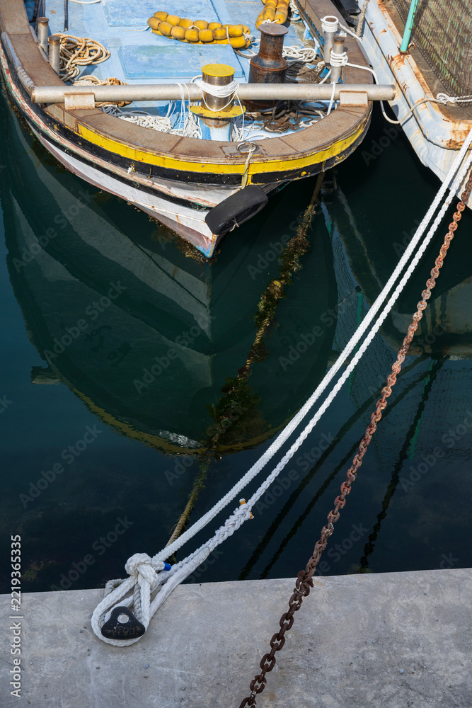 Fototapeta premium Small steel fishing boat moored at concrete pier with reflection on calm sea water tied by ropes and chain.