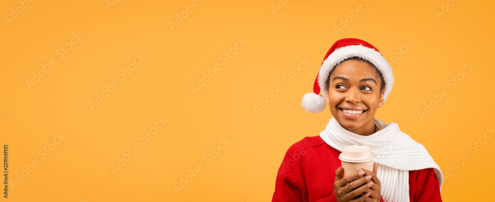 Fototapeta premium Happy Smiling Young Black Woman Wearing Santa Hat While Holding Coffee Cup, Standing Against Bright Yellow Background, Enjoying Holiday Spirit. Festive Concept For Christmas Celebrations.