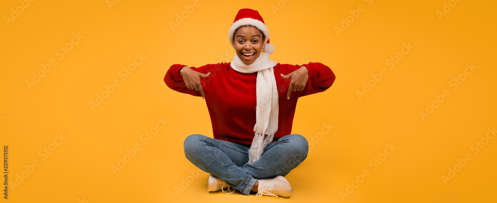 Fototapeta premium Smiling Young Black Woman In Santa Hat And Red Sweater, Sitting Cross-legged On Yellow Background, Pointing In Celebration, Expressing Joy And Happiness, Christmas Concept.