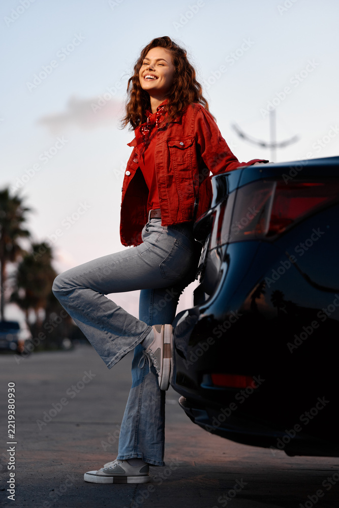 Fototapeta premium A happy young woman leaning against a black car during sunset. She wears a red jacket and blue jeans, smiling outdoors. Concept of freedom and travel lifestyle.
