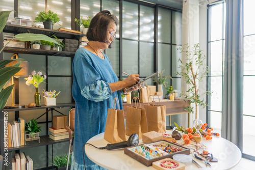 Mature Asian woman using tablet checking stock handcrafted jewelry at shop