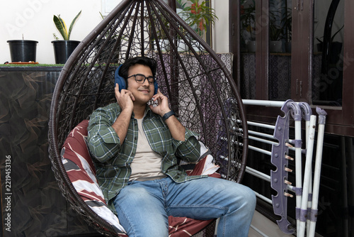Canvas Print Portrait of young handsome Indian man enjoying music with headphone while sitting on balcony at home