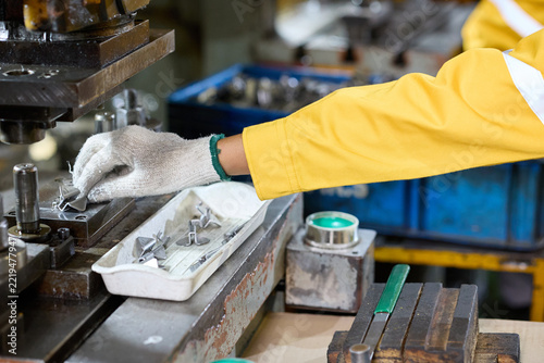 Worker's hand in a glove operating industrial machinery. The worker wears a yellow sleeve, and the scene shows manufacturing equipment and parts in a factory