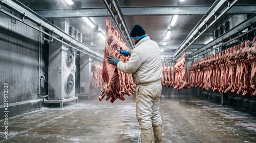 A worker in protective gear processes raw beef carcasses in a large industrial cold storage facility.