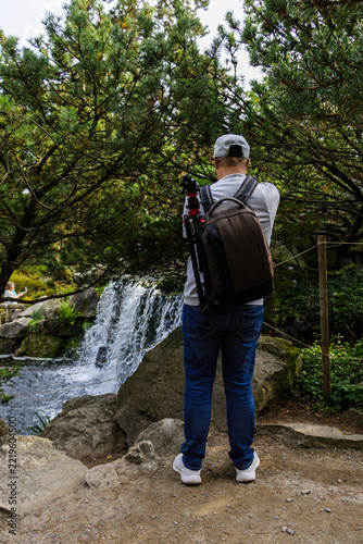 Photographer with Camera and Tripod Capturing Waterfall in Lush Green Garden