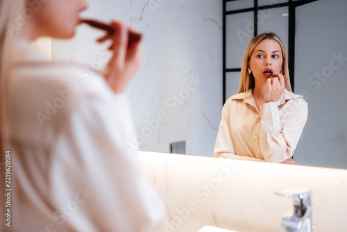 Woman checking her reflection while applying lipstick to her lips. Her reflected face is in focus as the cosmetic touches her mouth, with the shoulder and hand in front framing the mirror view.