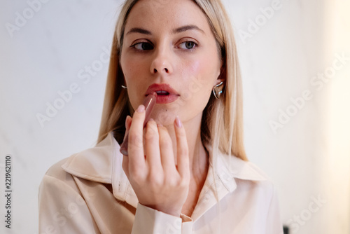 Woman carefully applying lipstick in a close facial portrait. Her fingers hold the cosmetic beside her mouth, emphasizing the lower lip, the gesture and her focused expression during grooming.