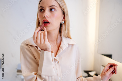 Woman using lipstick while looking to the side during her makeup. She holds the cosmetic at her lower lip with one hand, with a portrait showing her face, raised arm and attentive expression.