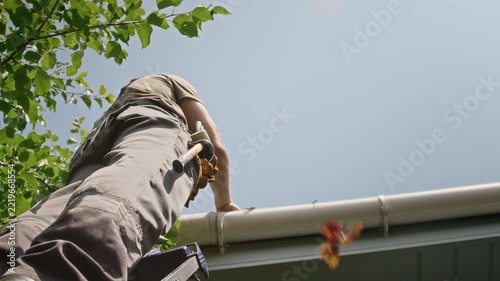 Male worker standing on ladder removing dry leaves from house roof gutter, low angle view. Ideal for residential maintenance advertisements and seasonal roofing service promotions