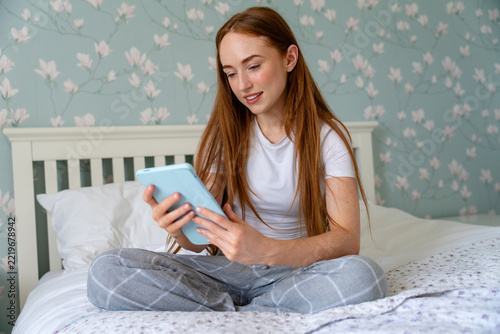 Girl sitting on a bed looking at a tablet while wearing casual clothes in a cozy room
