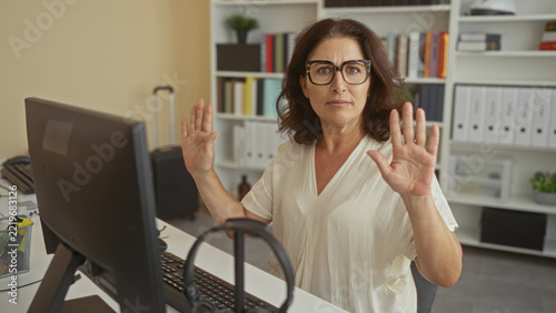 Fotografie Middle age woman hands raised palms out at computer desk in office with keyboard and monitor visible; defensive concern
