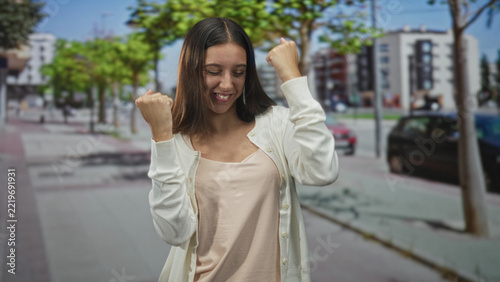 Fotografie Woman with raised fists celebrating on a city street wearing a cardigan and camisole; celebration success
