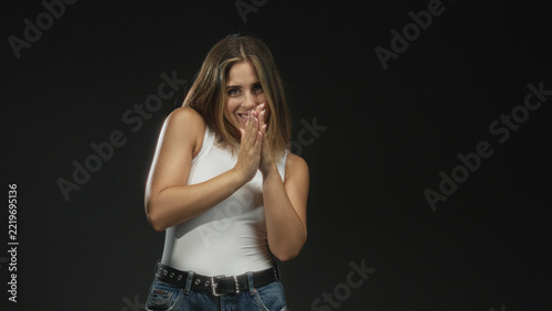 Fotografie Woman clasping hands near chest, smirking with coy gaze, wearing white tank and jeans in studio; playful anticipation