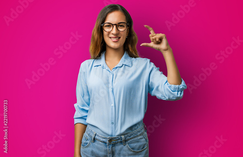 Young woman hispanic clothes in studio, confident and smiling, hand and fingers showing small size sign — finger gesturing to measure, small sign concept