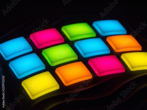 A close-up shot showcases a grid of brightly lit, colorful square buttons against a dark backdrop. The illuminated pads are in shades of blue, green, pink, orange, and yellow 