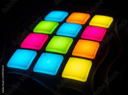 A close-up shot showcases a grid of twelve square pads, each illuminated with a different vibrant color, including blue, green, pink, orange, and yellow. The glowing pads are arranged in a 3x4 matrix 