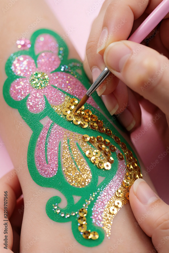 Naklejka premium Artist applies gold sequins to glitter flower tattoo on child arm. Pro makes temporary body art with pink sparkles, green patterns. Creative glitter makeup for kids at festive party birthday event