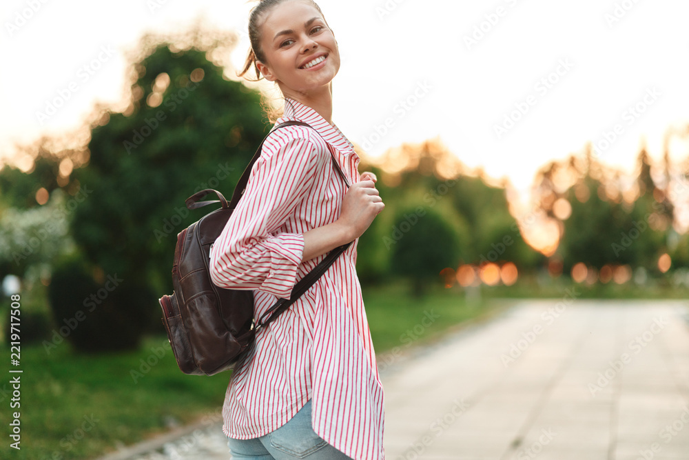 Fototapeta premium Young woman with a striped shirt and backpack smiles while walking along a sunlit path at dusk, conveying a carefree, hopeful mood and a sense of spontaneous everyday adventure.