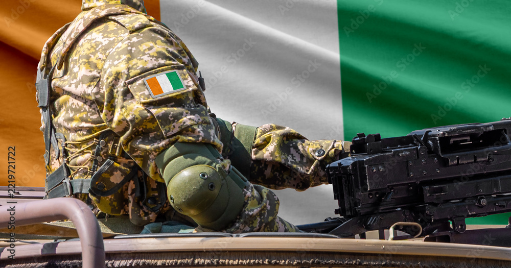 Fototapeta premium Ivory Coast flag with soldier at machine gun turret in front of it. Closeup side view.
