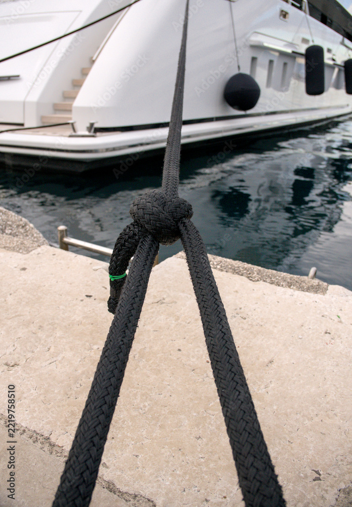 Fototapeta premium Black braided mooring rope knotted on quay with luxury yacht stern in blurred background.