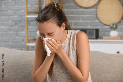 Cold and flu symptoms, young woman sneezing into tissue at home with seasonal...