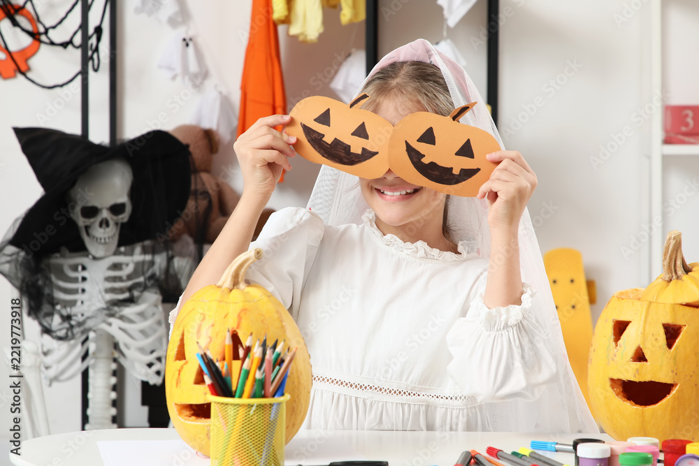 Fototapeta premium Halloween celebration and childhood creativity, happy girl wearing a bridal veil holding paper pumpkin masks over her eyes at home during autumn