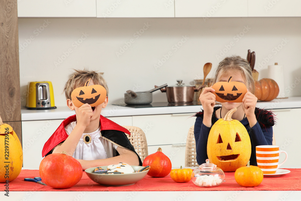 Fototapeta premium Halloween celebration and family fun, children in costumes holding paper pumpkin masks while sitting at a kitchen table with carved jack-o-lanterns