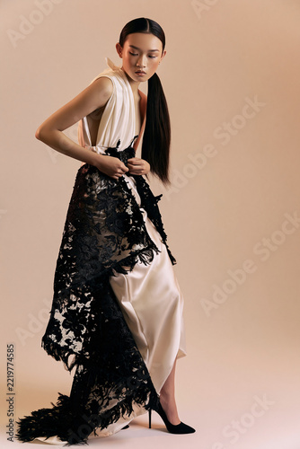 Elegant woman adjusting black floral lace over an ivory couture gown against a warm beige studio backdrop