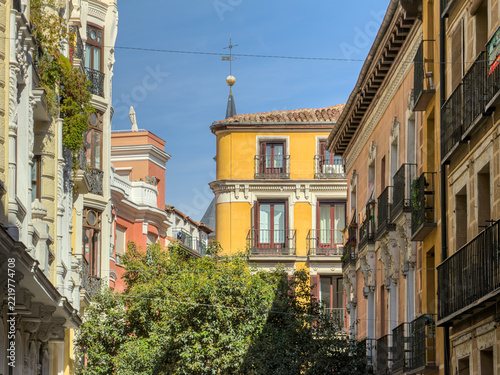 Charming traditional residential buildings with ornate balconies and colorful yellow facades on a sunny street in Madrid, Spain, surrounded by lush green trees.