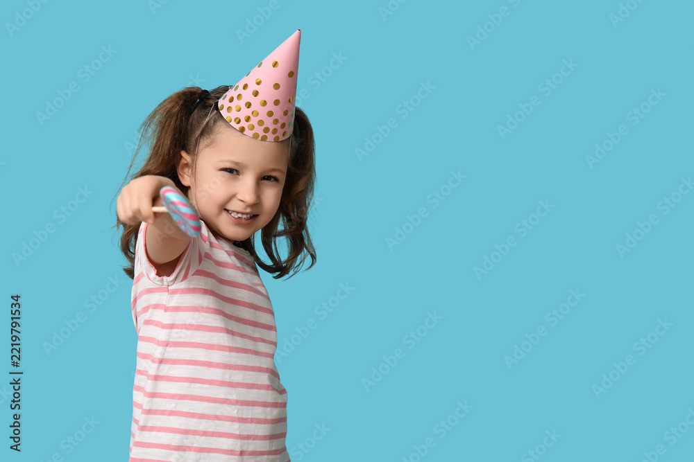 Fototapeta premium Birthday celebration and childhood joy, happy little girl wearing a party hat holding a colorful lollipop on a solid blue background