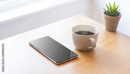 A smartphone and a cup of coffee sit on a wooden desk near a plant.
