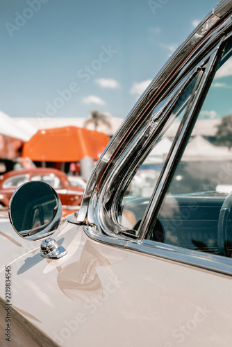 Classic vintage car detail featuring polished chrome trim, side mirror, and white bodywork under a bright blue sky.