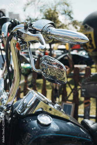 Ornate chrome motorcycle handlebars and engraved mirror at a vintage motorcycle exhibition under bright daylight.