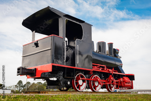 Historic black and red steam locomotive displayed outdoors on railway tracks beneath a dramatic blue and white sky.