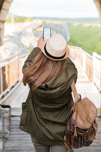 Rear View of Female Traveler with Backpack Taking Photo with Smartphone on Scenic Wooden Bridge