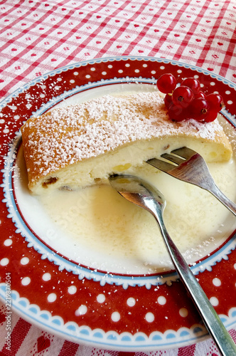 A slice of traditional Bavarian quark strudel dusted with powdered sugar, served with creamy sauce and red currants on a decorative red plate