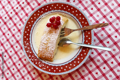 A slice of traditional Bavarian quark strudel dusted with powdered sugar, served with creamy sauce and red currants on a decorative red plate