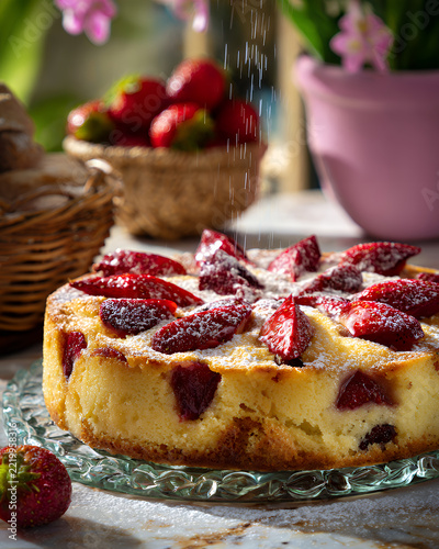 Fresh baked strawberry cake on a glass plate being dusted with powdered sugar with a basket of berries