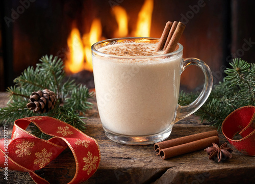 Glass mug of creamy eggnog with cinnamon sticks and pine branches near a warm fireplace