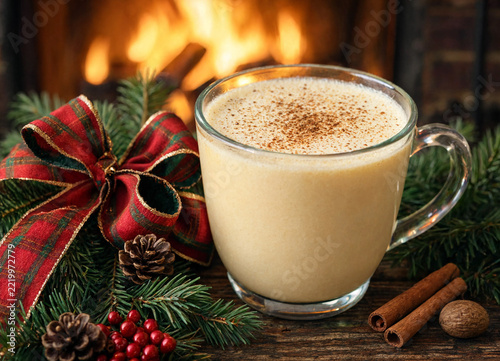 Glass mug of eggnog with Christmas decorations and a cozy fireplace in the background