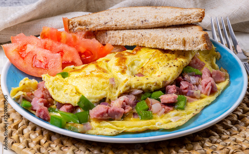 A breakfast Denver omelette filled with ham, green bell pepper and onion with tomato slices and while grain toast bread