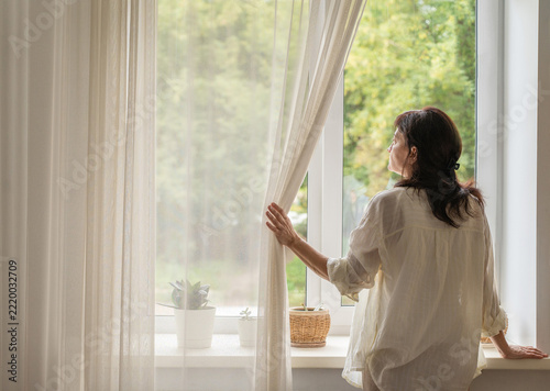 Middle-aged woman stands with her back to camera by window, parting light curtains and gazing at trees, immersed in morning reflection. Slow living, mindful lifestyle after 50