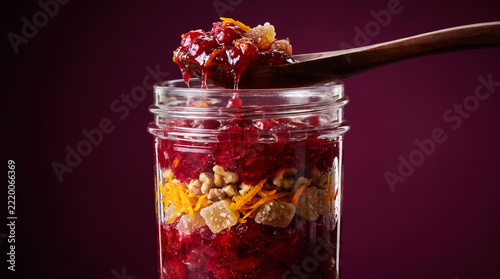 Clear Glass Jar of Homemade Cranberry Orange Relish with Walnuts, Candied Ginger and Wooden Spoon Against a Deep Burgundy Background